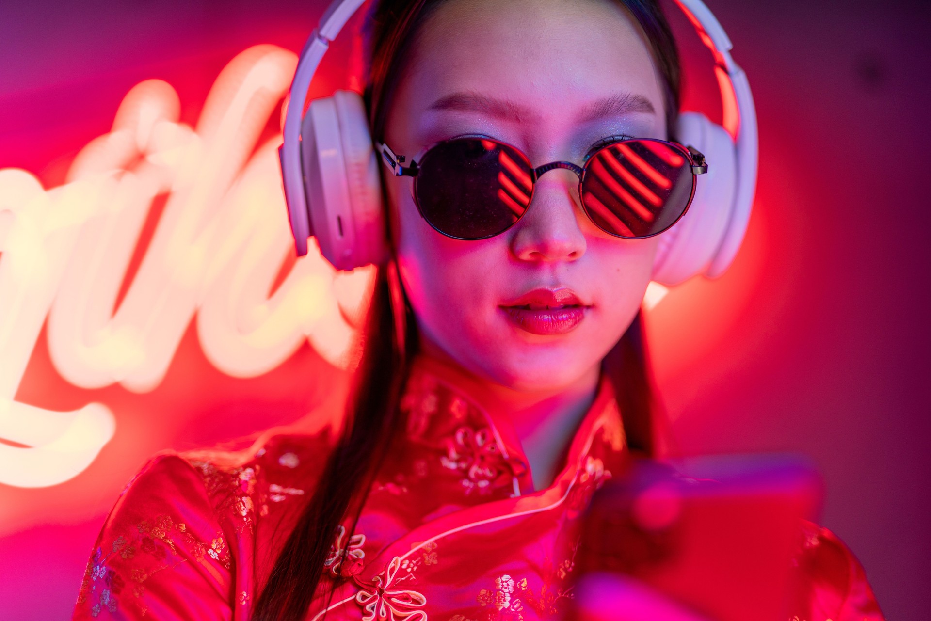 Close up portrait photo of a beautiful young asian female lady model with cool look wearing sunglasses, earphone and red traditional chinese cheongsam dress in front of orange pink neon lights
